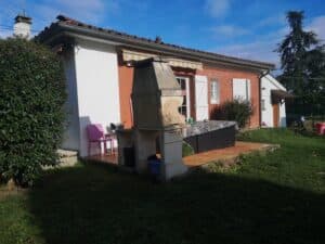 Maison de plain-pied avec façade brique/blanche, barbecue en béton, salon de jardin sur terrasse carrelée, et jardin verdoyant sous un ciel bleu.