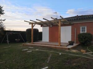 Construction d'une pergola en bois sur une terrasse attenante à une maison en briques rouges, avec une balançoire dans le jardin.
