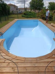 Installation d'une nouvelle piscine avec revêtement bleu clair et terrasse en bois, un homme surveille les travaux dans un jardin.