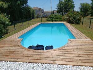Piscine hors-sol avec terrasse en bois clair, entourée de verdure et de gravier blanc, sous un ciel bleu.